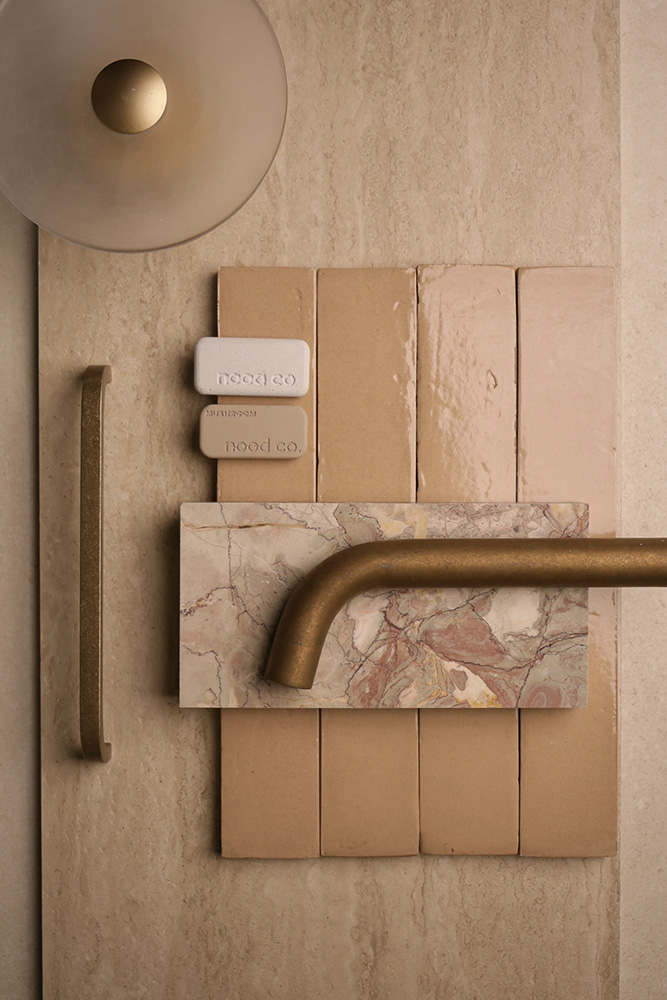 A palette flatlay on a travertine surface — Zellige Look Feature Wall Tile in natural, an Italian-made ceramic look-alike laid in slim 50×150mm vertical format with the soft hand-touched glaze and natural variation of real zellige. Paired with a brushed brass linear cabinet handle, a smoked glass dome wall sconce with brass ball, a brushed brass wall-mounted basin spout, two Nood Co. bathware sample chips in cream and mushroom, and a sage-and-blush veined marble vanity sample. A complete palette curated by the Design Tiles team at our Rockdale showroom.