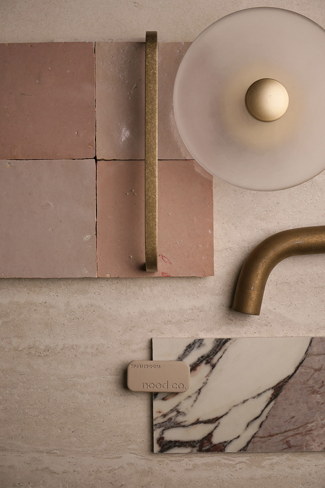 A second palette flatlay on travertine — Zellige Moroccan Tile Handmade Glazed in blush, authentic Fez-made zellige with raw clay edges and hand-poured glaze variation. Paired with a brushed brass cabinet handle, a smoked glass dome wall sconce, a brushed brass basin spout, a Nood Co. mushroom-toned bathware sample, and a Calacatta Viola marble vanity sample with dramatic burgundy veining. A second palette direction curated by the Design Tiles team at our Rockdale showroom.