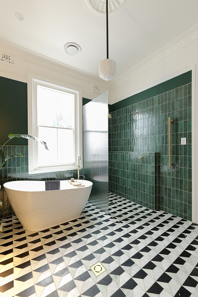A Sydney ensuite with vertical forest green handmade Moroccan zellige wrapping the walk-in shower and bath alcove, a freestanding white bath beneath a tall sash window, fluted glass shower screen, brushed brass shower fittings and towel rail, ornate plaster ceiling rose, period pendant light, and a black-and-white marble cubed tile floor. The handcrafted glaze catches the natural daylight unevenly across each tile.