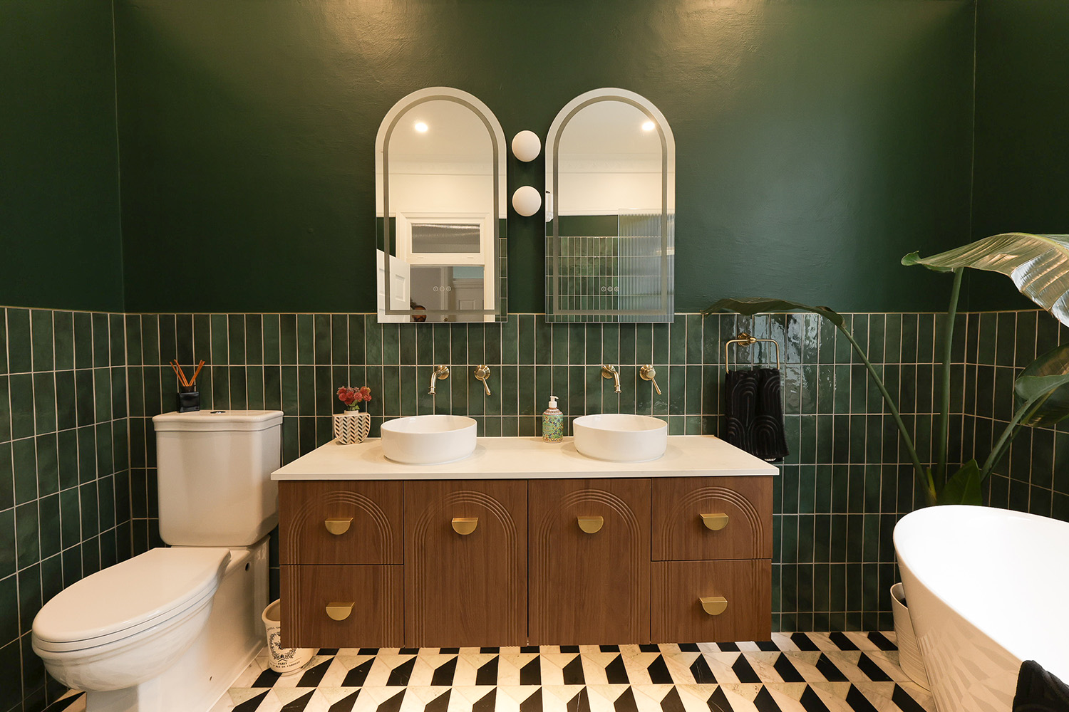 A Sydney bathroom featuring a deep forest green handmade Moroccan zellige feature wall laid in vertical stack bond, with twin arched mirrors and white globe sconces above a dark walnut fluted vanity with brass half-moon handles, white vessel basins on a stone benchtop, brushed brass wall-mounted tapware, and a dramatic black-white-and-grey geometric cubed marble floor. Zellige used as a confident colour statement in a moody, design-led Sydney ensuite.
