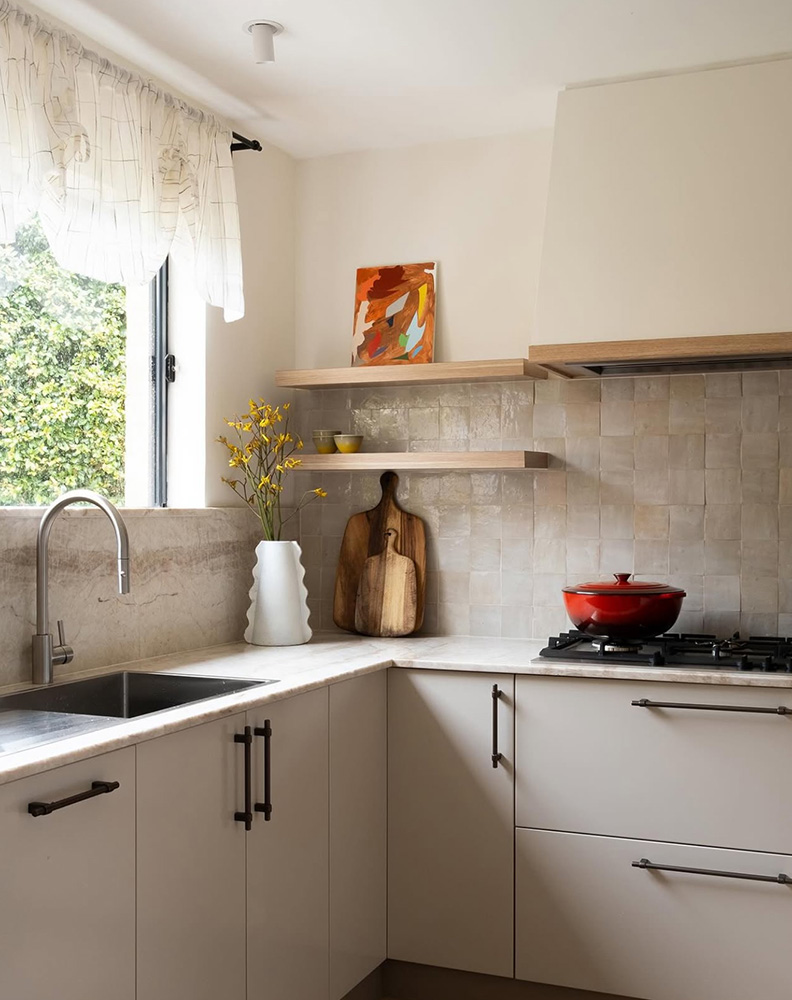 A Sydney kitchen with a soft natural handmade Moroccan zellige backsplash in 100×100mm squares, paired with a marble waterfall benchtop, light timber open shelving, cream cabinetry with bronze hardware, a sheer linen curtain at the window, and a red Le Creuset on the gas cooktop. Zellige used as a soft, sun-warmed kitchen anchor in a refined Australian interior.